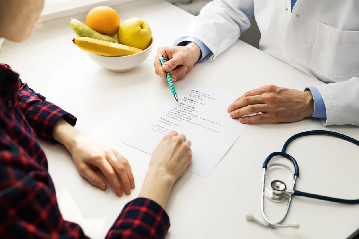 Two people sit at a table with a document, fruits in a bowl, and a stethoscope. One points at the paper with a pen.